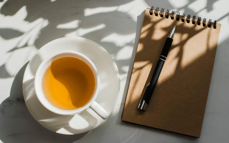 Cup of herbal green tea at marble table with notebook. Hard sun light and plant shadows